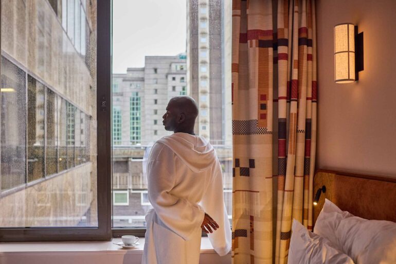 Folu Odimayo wears a linen robe while looking out over rainy London from his hotel room at the Sir Devonshire Square Hotel