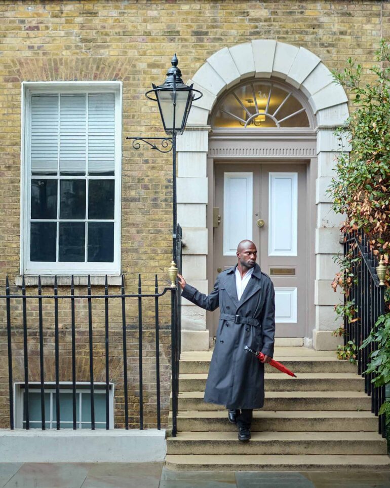 Folu Odimayo descending a stairway wearing a trench coat and a red umbrella in London, UK