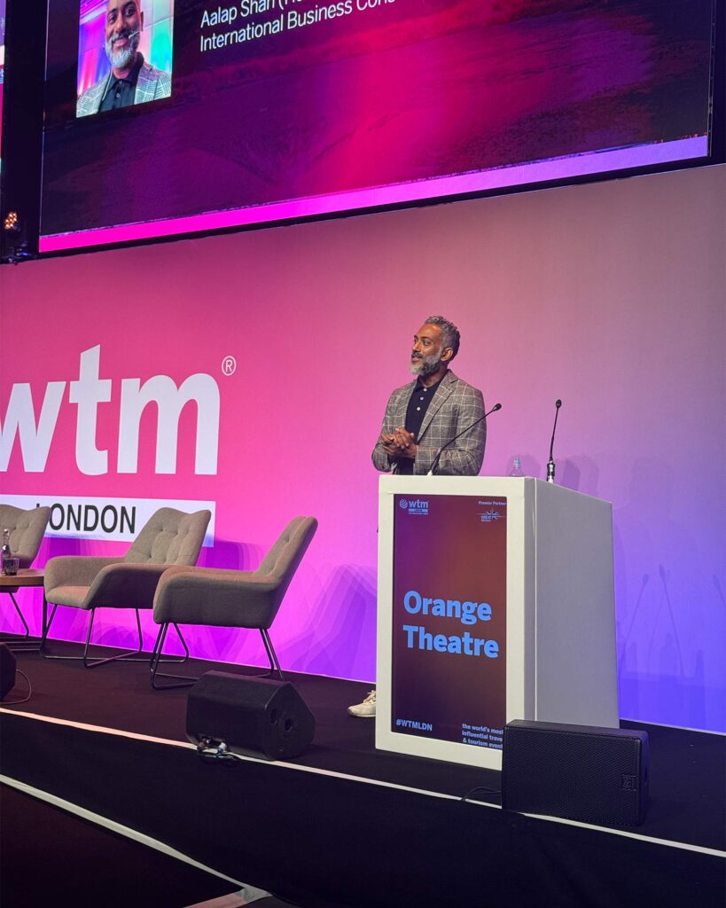 A speaker stands behind a podium marked “Orange Theatre” at WTM London, addressing the audience with a large screen above displaying their photo and name.
