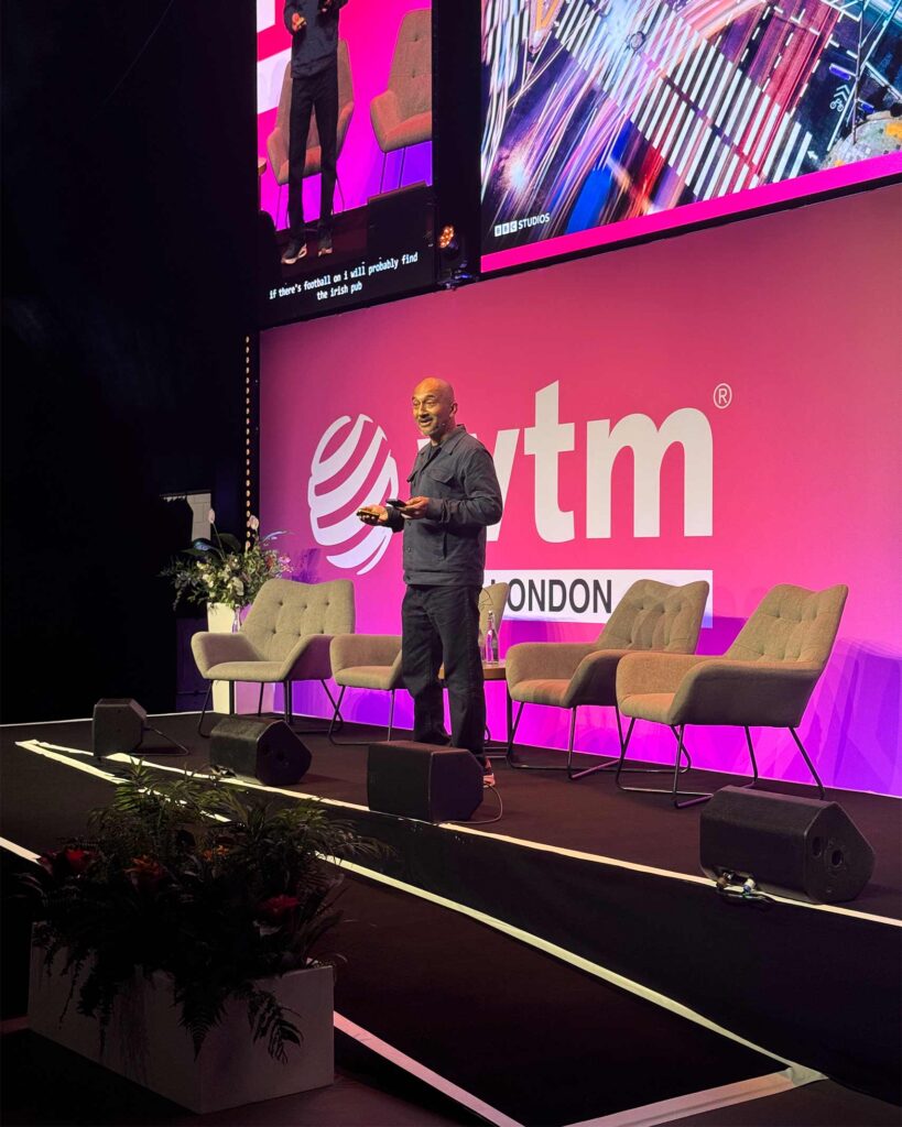 A solo speaker stands mid-presentation on stage at WTM London’s Orange Theatre, holding a device and gesturing toward the audience, with a large visual display projected overhead.