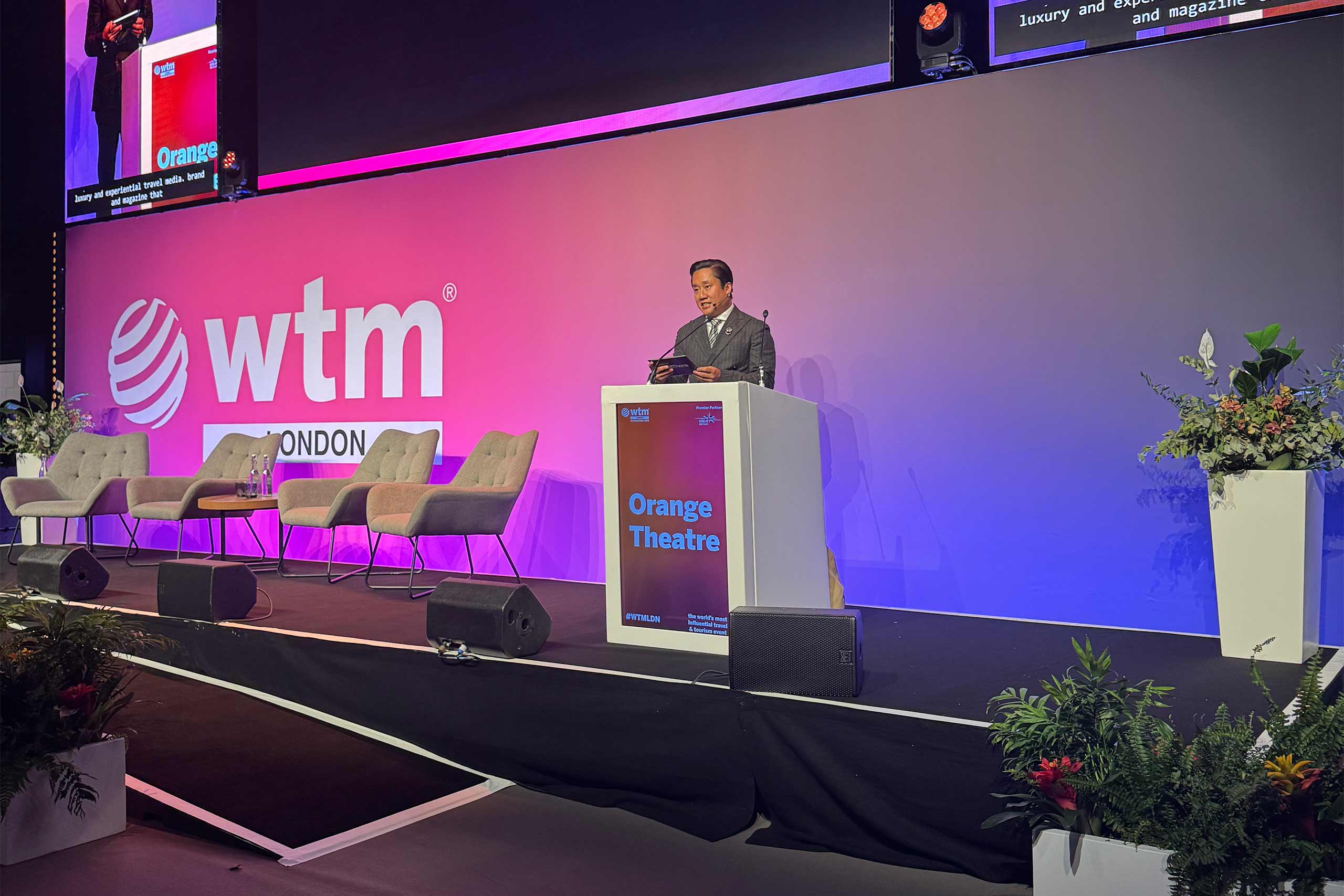 A presenter stands behind a podium marked “Orange Theatre”, speaking to an audience. Several empty chairs and the event’s bright pink backdrop with the event logo are visible behind them.