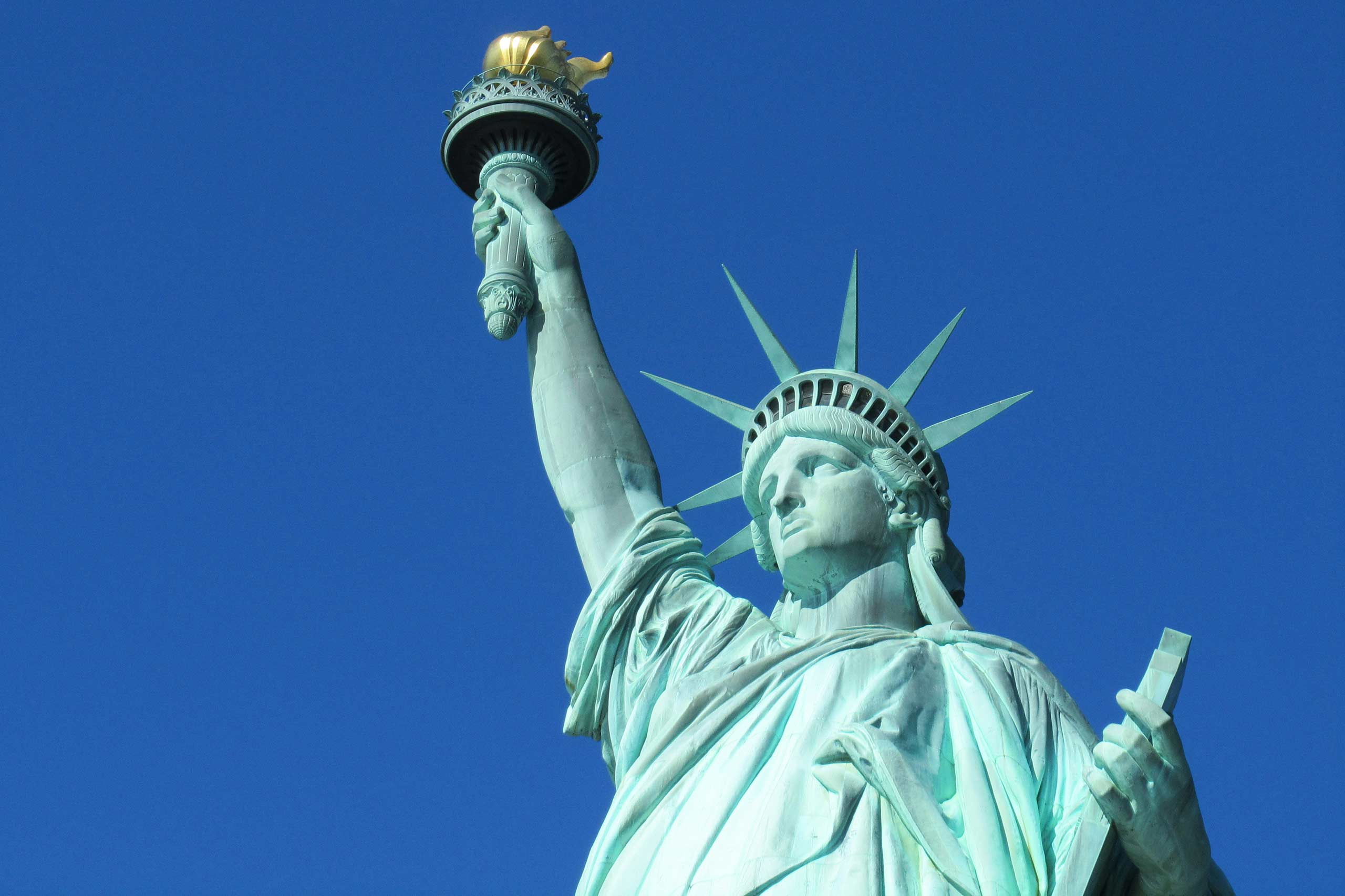 The top half of the statue of liberty, green to a backdrop of a crisp blue sky, illustrating the state of the politics of travel today