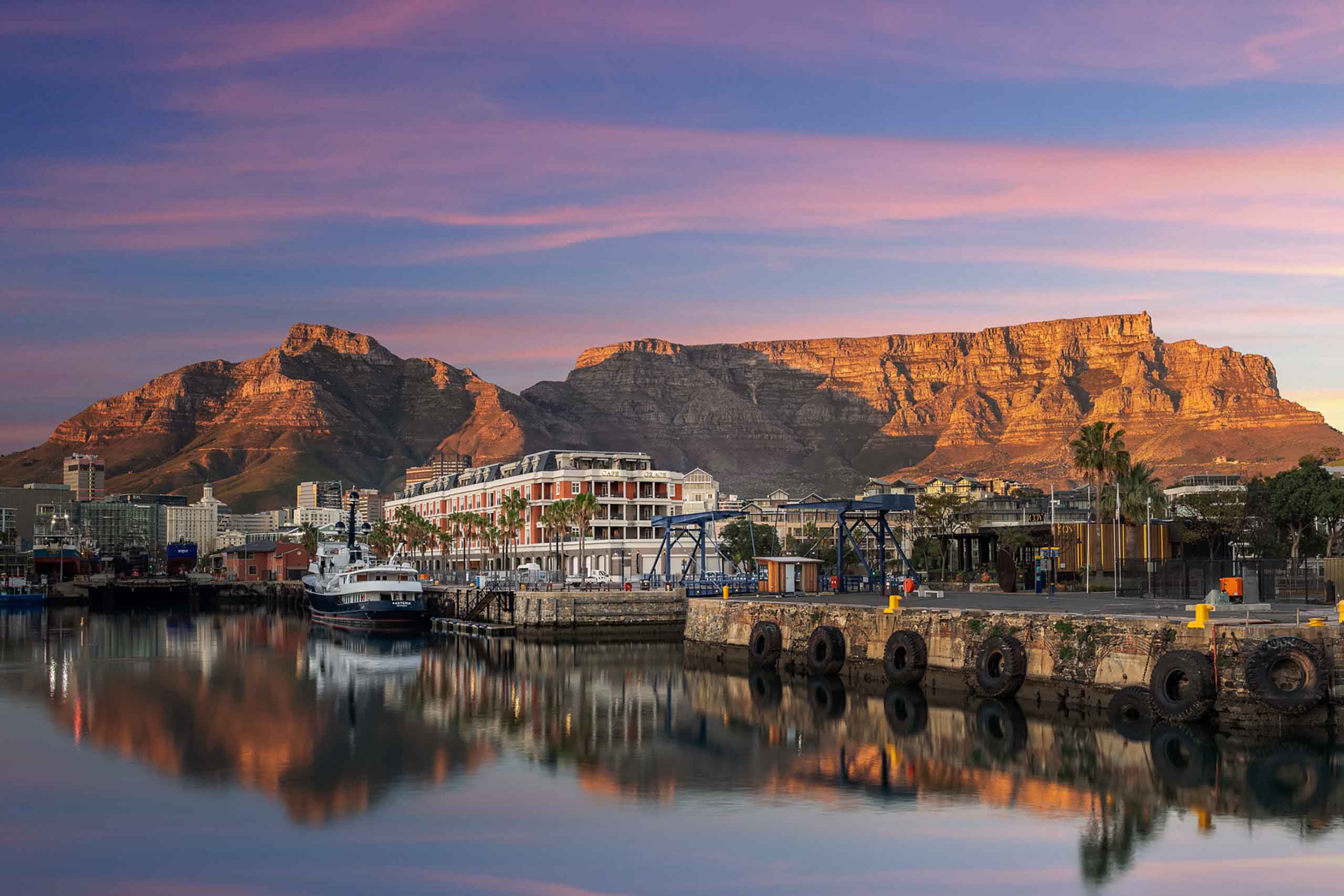 Exterior shot of Cape Grace hotel from the V&A Waterfront at dusk, depicting the old building in the mid-ground, a sunset hued Table Mountain in the background and stunning light reflected in the water in the foreground