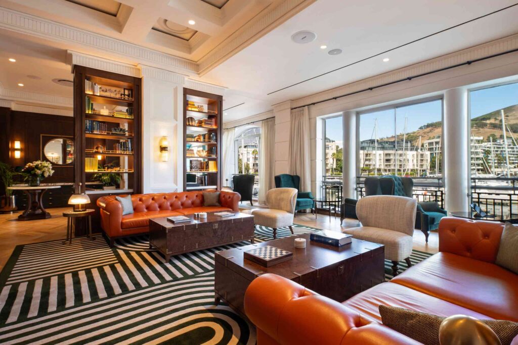 Library lounge at Cape Grace overlooking the marina. The interiors feature orange sofas, blue patterned carpets, wooden trunks and shelves full of books and objets d'art