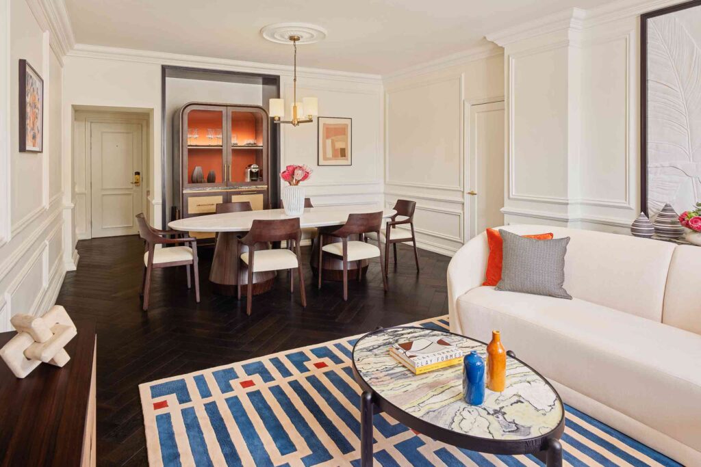 Suite living room interiors, featuring a large stone dining table with wooden chairs, armoire minibar, dark wooden floors, blue, red and white carpet and white sofa