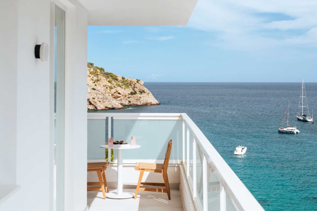 View from a white hotel balcony overlooking a craggy headland and blue sea with yachts