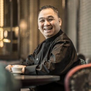 Portrait of Jimmy Wong, Executive Chef, Four Seasons Kuala Lumpur. Jimmy is a Chinese man, wearing a black chef's tunic with a mandarin collar. His hands are placed by a white bone china chinese teacup. He is smiling