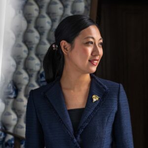 Portrait of Melody Wong, Director of Wine at Peninsula London. Melody is a Chinese woman, has black hair tied back in a ponytail and she wears a blue suit. On her lapel is a 'bunch of grapes' pin, indicating she is a sommelier