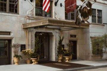 Exterior with front entrance detail of The Surrey, a Corinthia hotel in New York City. Plants adorn its 1920 doorway and a brown carpet with a monogram S, covered by an awning with the hotel name on it
