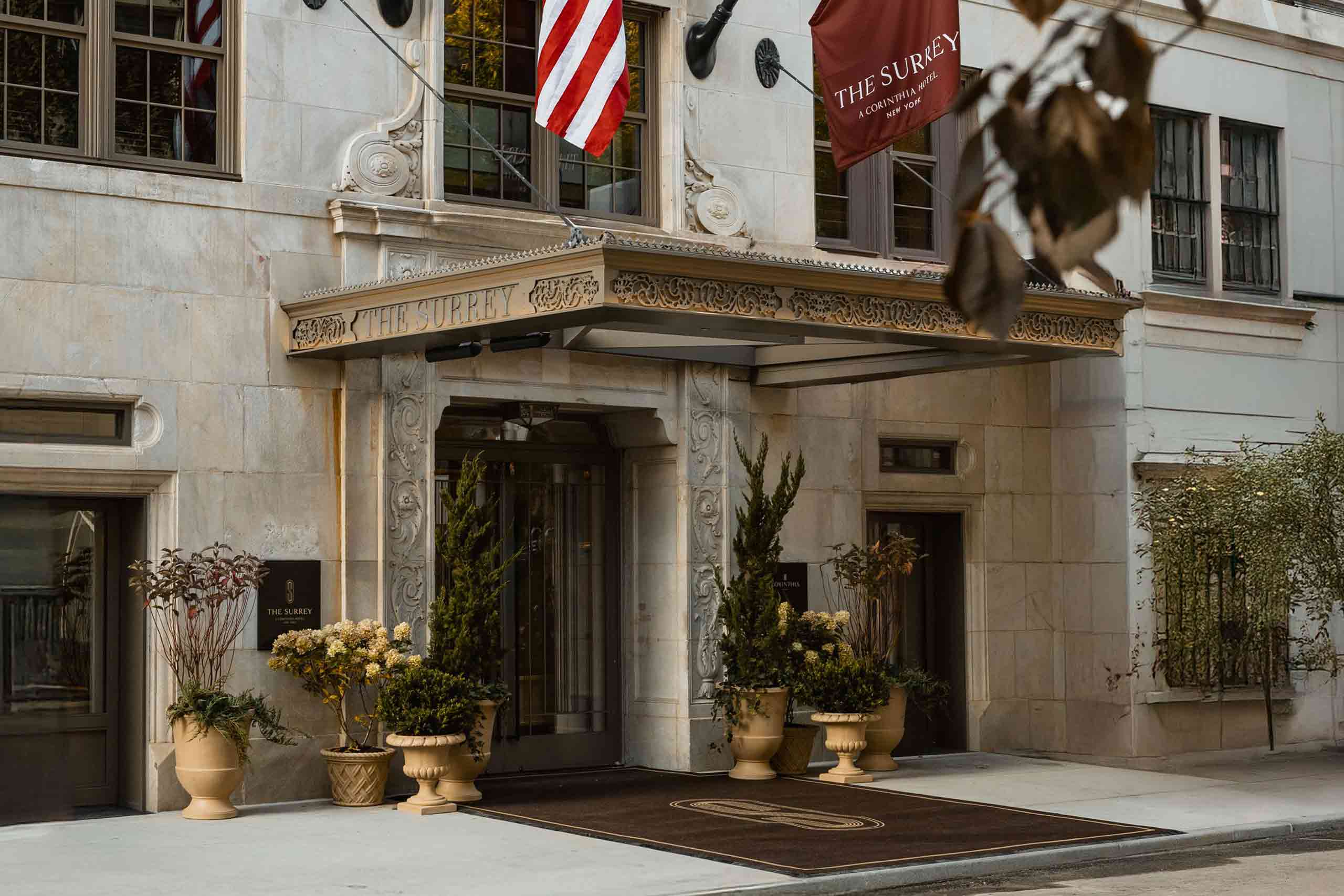 Exterior with front entrance detail of The Surrey, a Corinthia hotel in New York City. Plants adorn its 1920 doorway and a brown carpet with a monogram S, covered by an awning with the hotel name on it