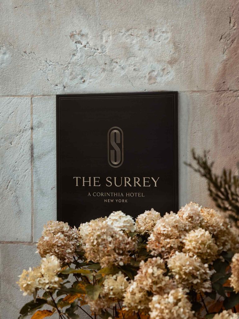 Brown entry sign on the building's white stonework that bears The Surrey's monogram S and reads "The Surrey, a Corinthia Hotel, New York". Below it are flowers that partly cover the sign