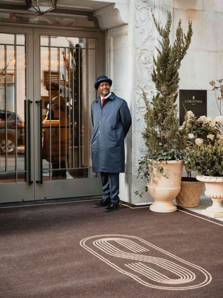 A black doorman stands at the entrance of The Surrey with his hands behind his back. He smiles. He is wearing a blue bowler hat and a blue trenchcoat over his uniform white shirt, red tie and blue trousers
