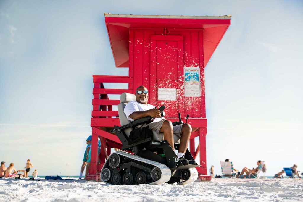 A male wheelchair user poses for a picture on the beach in his beach-adapted wheelchair