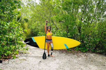 An amputated female paddle boarder poses with a yellow paddle board in front of lush vegetation; an example of what accessible Florida looks like