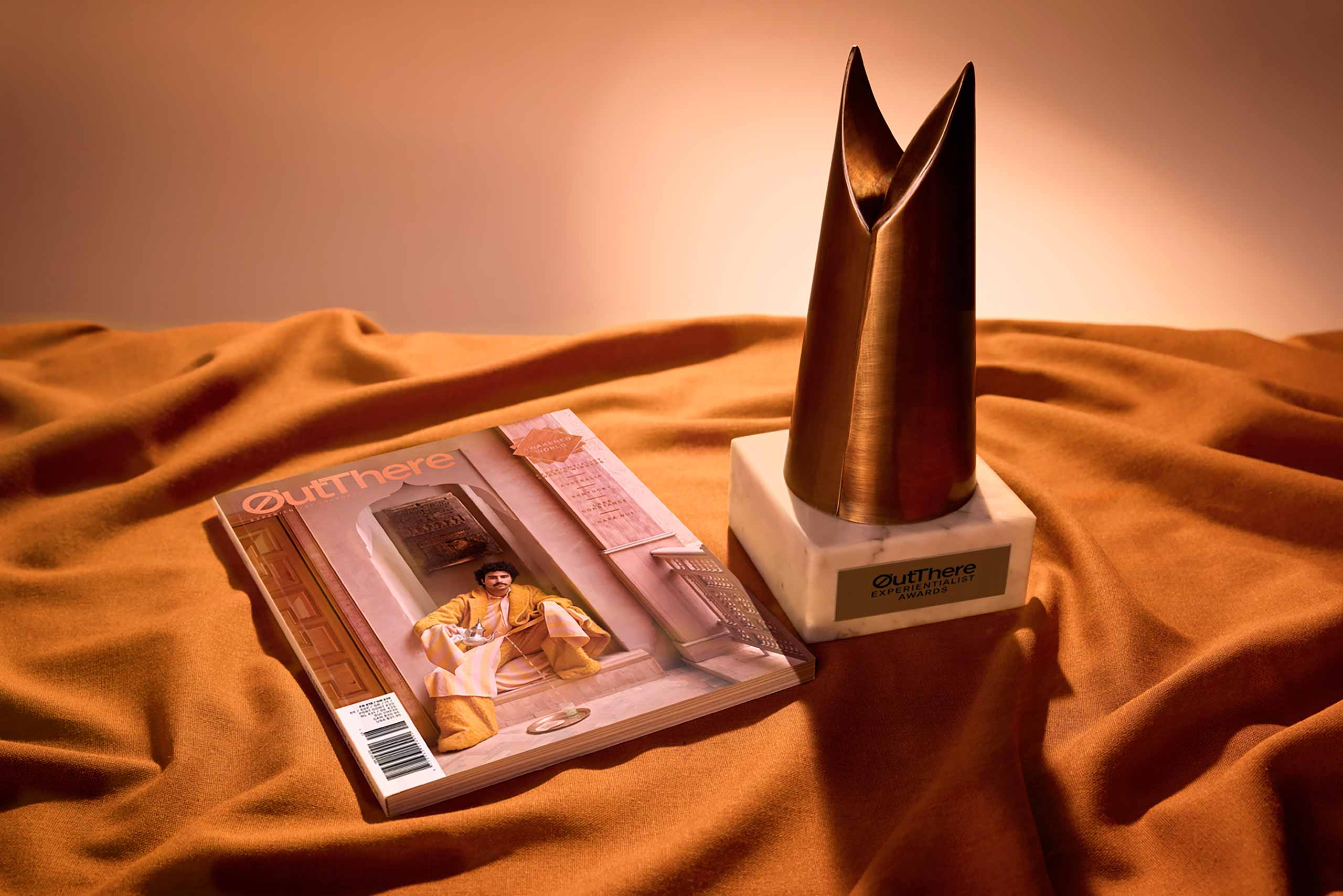 Experientialist® Awards 2026 by OutThere, hand-forged copper trophy and our Awakend World Issue photographed on copper coloured satin cloth