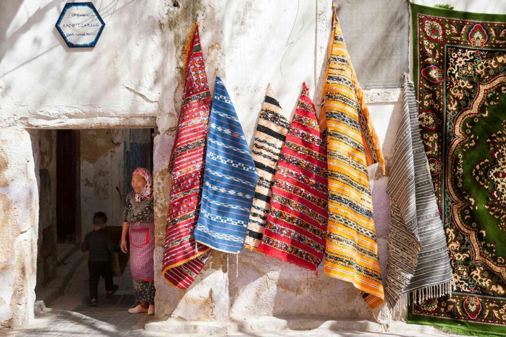 A woman peeks out from a white souk doorway, while colourful carpets hang from the wall in Fes, Morocco
