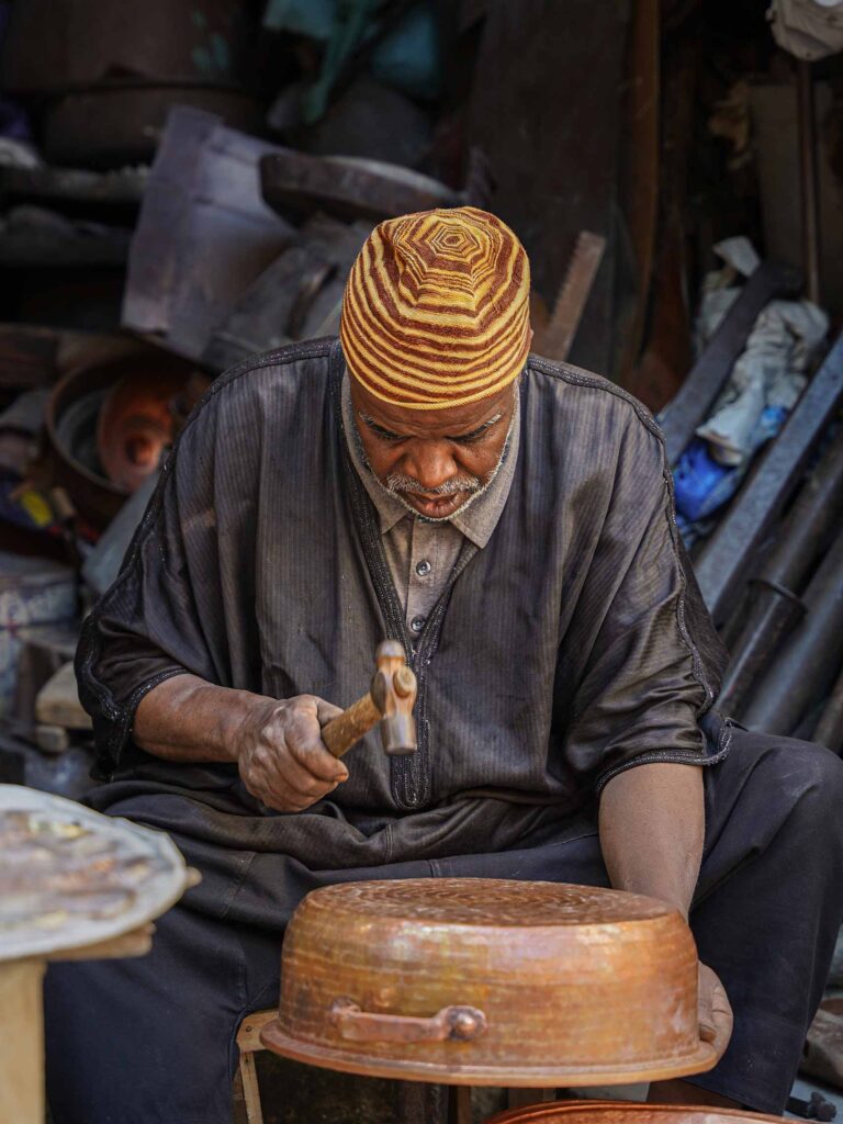 A craftsman in Fes wears a grey robe and yellow hat as he hammers a copper pot