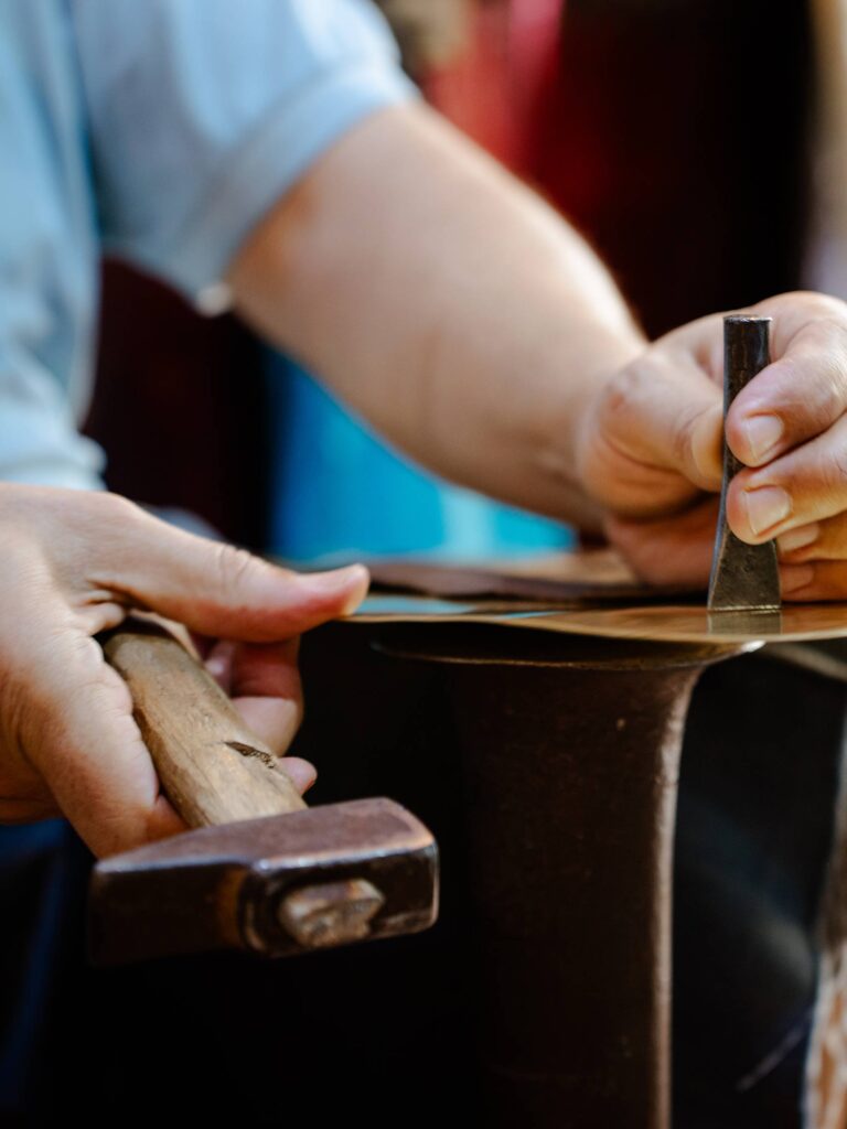 Arms and hands of a Moroccan artisan in a blue shirt as he hammers, engraves and shapes the flat copper into a piece of art