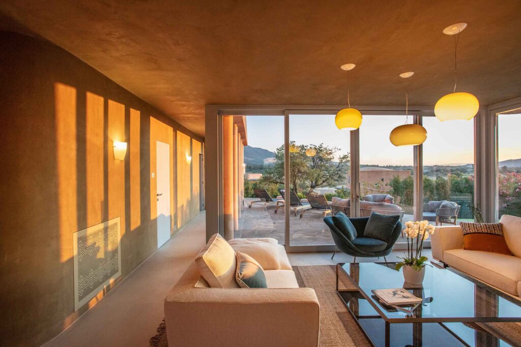 Interiors of Villas at Is Molas, Sardinia – terracota walls, peach sofa, blue armchair, Italo-chic glass ball light fittings looking out through floor to ceiling windows to the garden