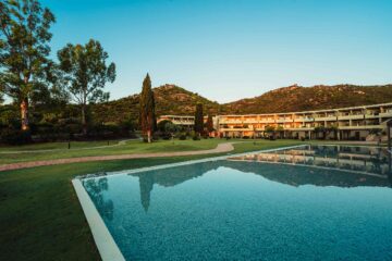 View of Is Molas Resort, overlooking its palm fringed, irregularly shaped, turquoise blue pool and to its curved mid-century-style main building set in lush greenery