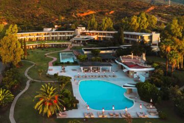 Aerial view of Is Molas Resort, overlooking its palm fringed, irregularly shaped, turquoise blue pool and to its curved mid-century-style main building set in lush greenery