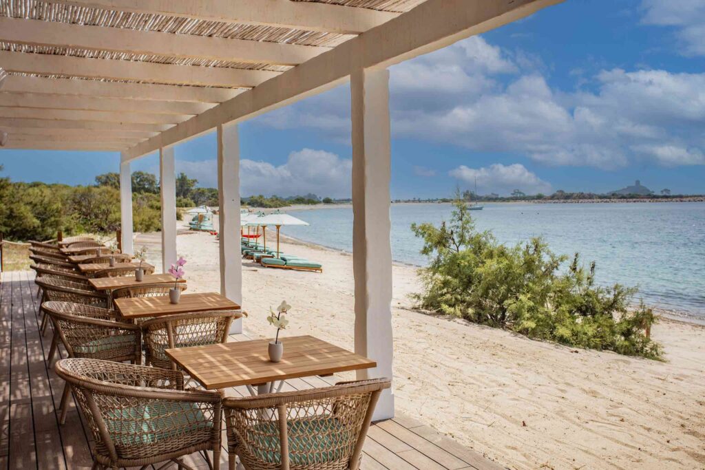Is Molas Resort Sardinia beach club restaurant, rattan chairs on a terrace look out to the sandy beach and water. The beach is rugged with shrubs and where clear, turquoise blue sun loungers sit under turquoise and white striped sunloungers