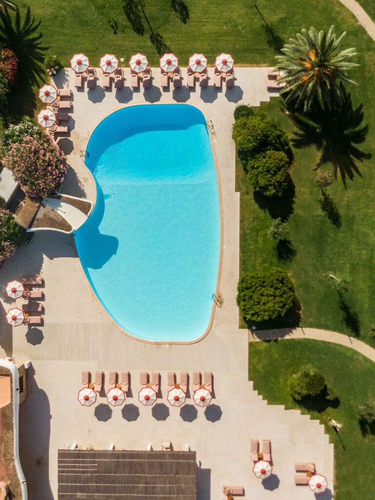 Drone shot of the pool deck at Is Molas Resort, overlooking its palm fringed, irregularly shaped, turquoise blue pool, flanked by terracotta sun loungers and red and white parasols