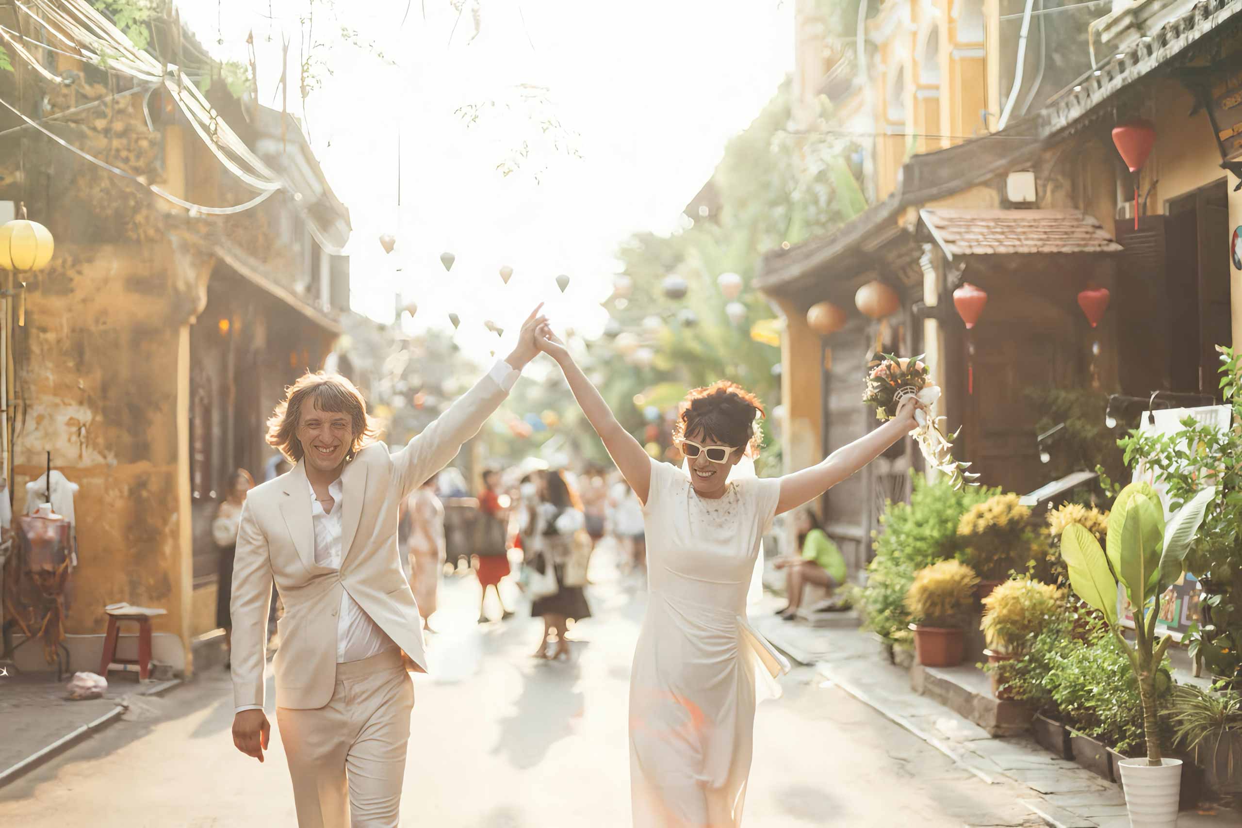 Two women, one in a white suit, the other in a white dress, sunglasses and bouquet, hold their hands up together on a street in Hoi Ann Vietnam. The image insinuates they're recently wed, and is an example of milestone travel