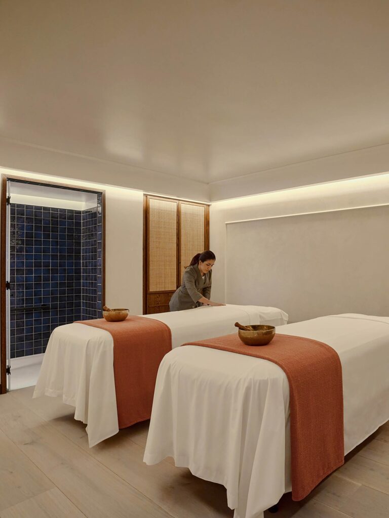Spa therapy room at Six Senses London, a therapists shoots a white spa bed. On each of them, orange towels and a brass healing sound bowl