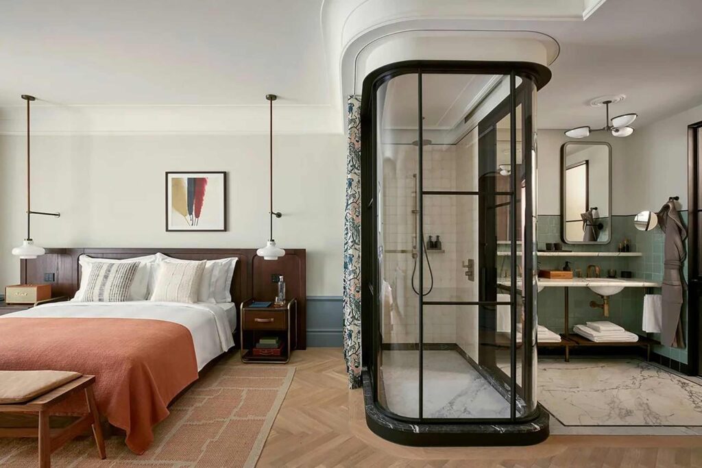 Room at Six Senses London with wood headboard bed, art deco lights, orange throw, open plan bathroom and glass encased wet-room shower