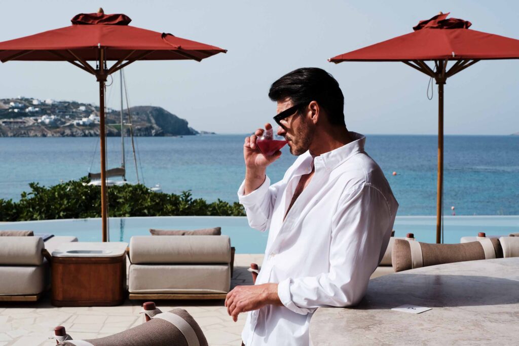 A dark haired man in a white linen shirt leans on the bar and drinks a red cocktail, with an infinity pool overlooking the sea in Greece. He is flanked by two burnt sienna parasols