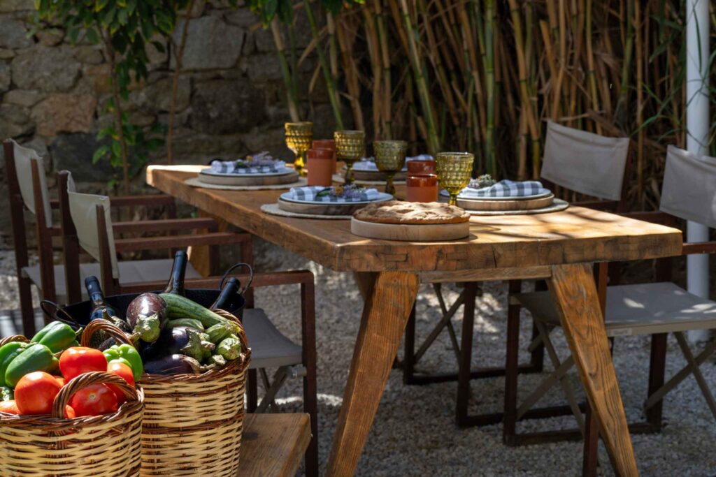 A rustic, designer, wooden dining table is set while fresh vegetables in baskets and bottles of wine sit in the foreground