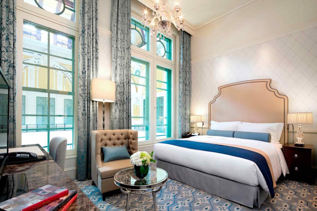 Bedroom at the Tokyo Station Hotel being flooded with light through two large windows. In the room, a king sized bed with big headboard, arm chair, crystal chandelier, desk and patterned carpet
