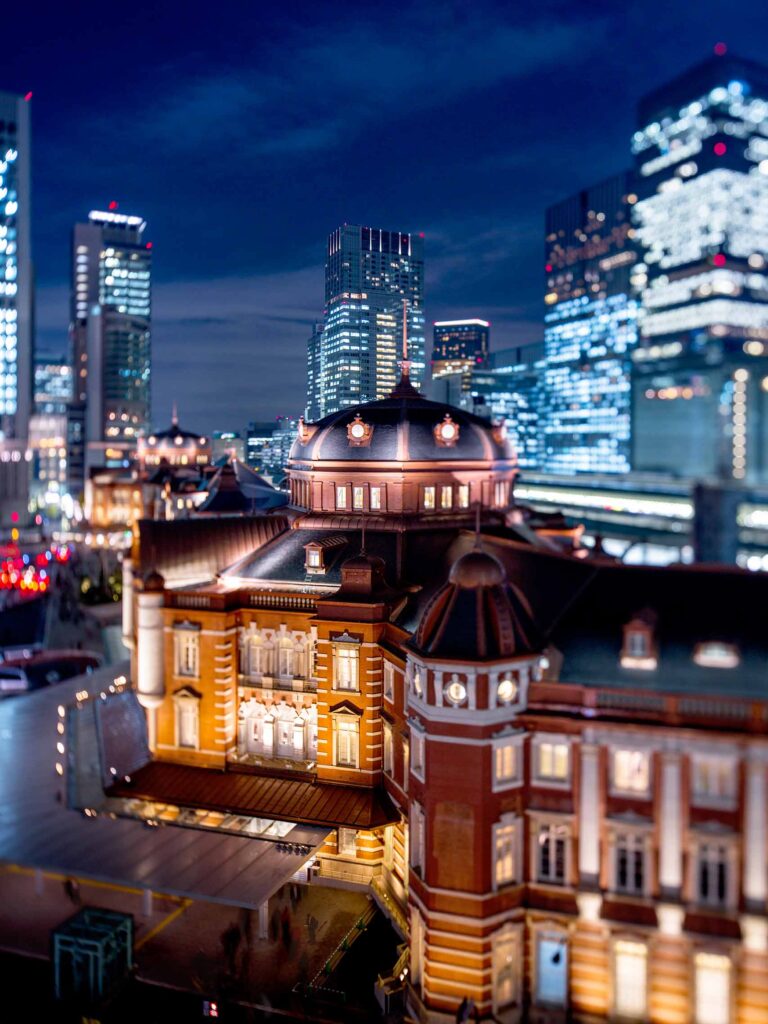 Exterior shot of the red-brick, domed roof of the Gothic Revival architecture The Tokyo Station Hotel, set to a backdrop of tall, glass skycrapers in Japan at night