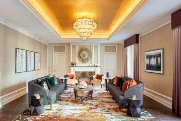 Two large sofas in front of a fireplace and beneath a golden ceiling from which a chandelier hangs at Fairmont Hotel Vancouver, Canada