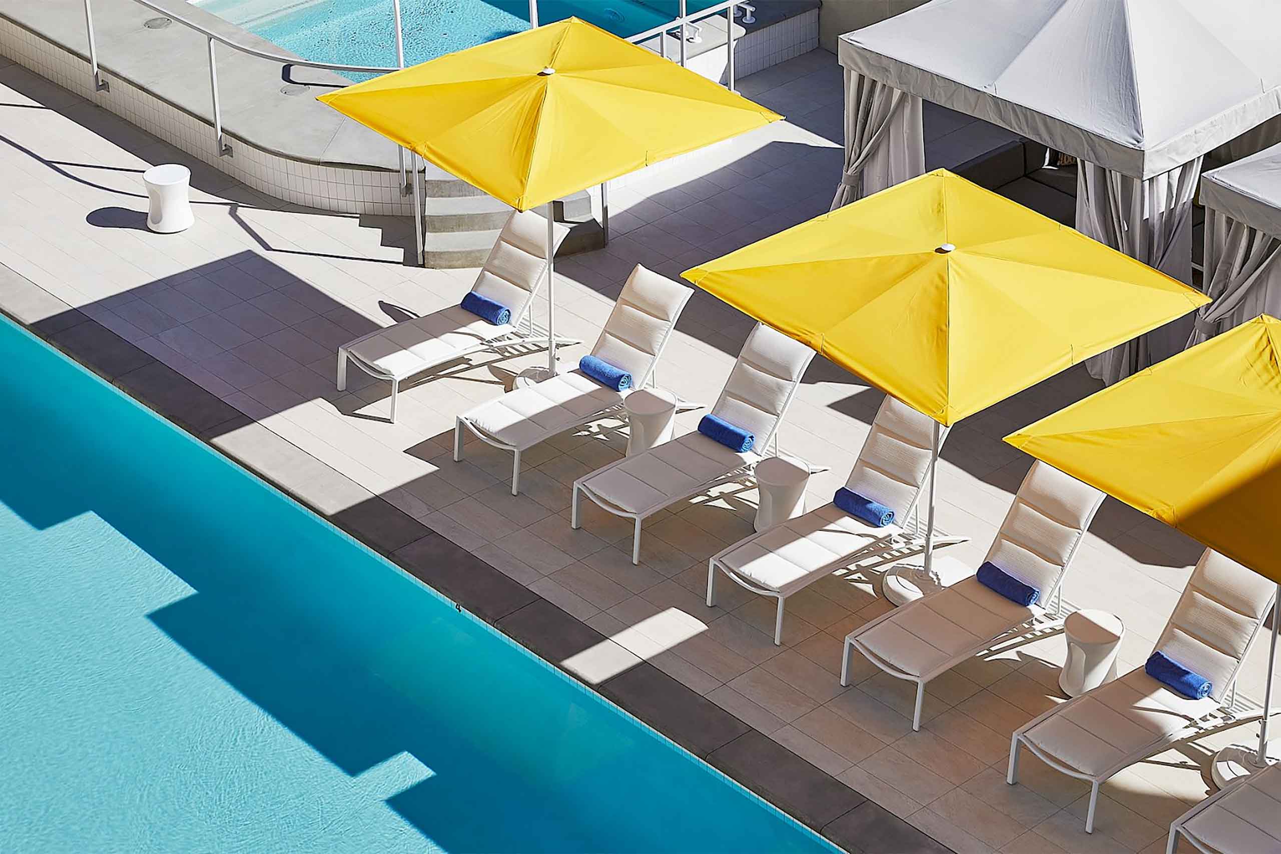 A sunny poolside lined with white sun-loungers and rolled-up blue towels, with yellow parasols