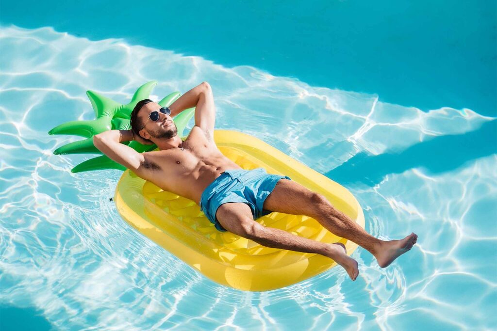 A white man with a beard, sunglasses and blue swim shorts lies back on a pineapple shaped float in the pool of Hotel Paseo