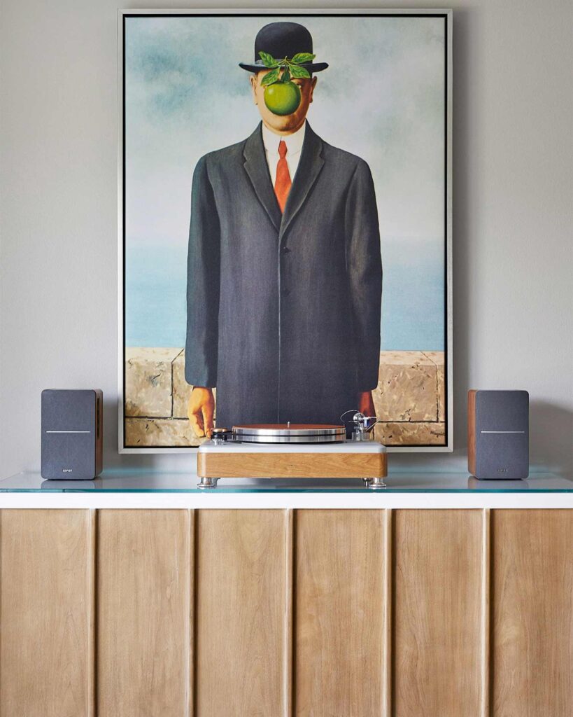 A record player sits on a beech sideboard with speakers either side. Above it a piece of art depicting a man in a suit and bowler hat, his face obscured by a green apple