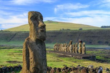 A moai stone statue on Rapa Nui, with head and big nose carved on Easter Island in Chile. Behind it a row of further moai, 15 in total all standing like guardians on the green landscape