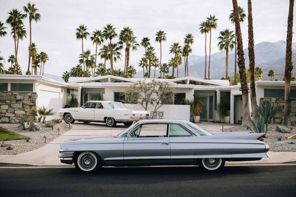 A silver 1950s vintage car parks outside a white Mid Century Modern bungalow in Palm Springs. The the background, tall palm trees and the mountain feature. Parked in the house's porch is a white vintage car