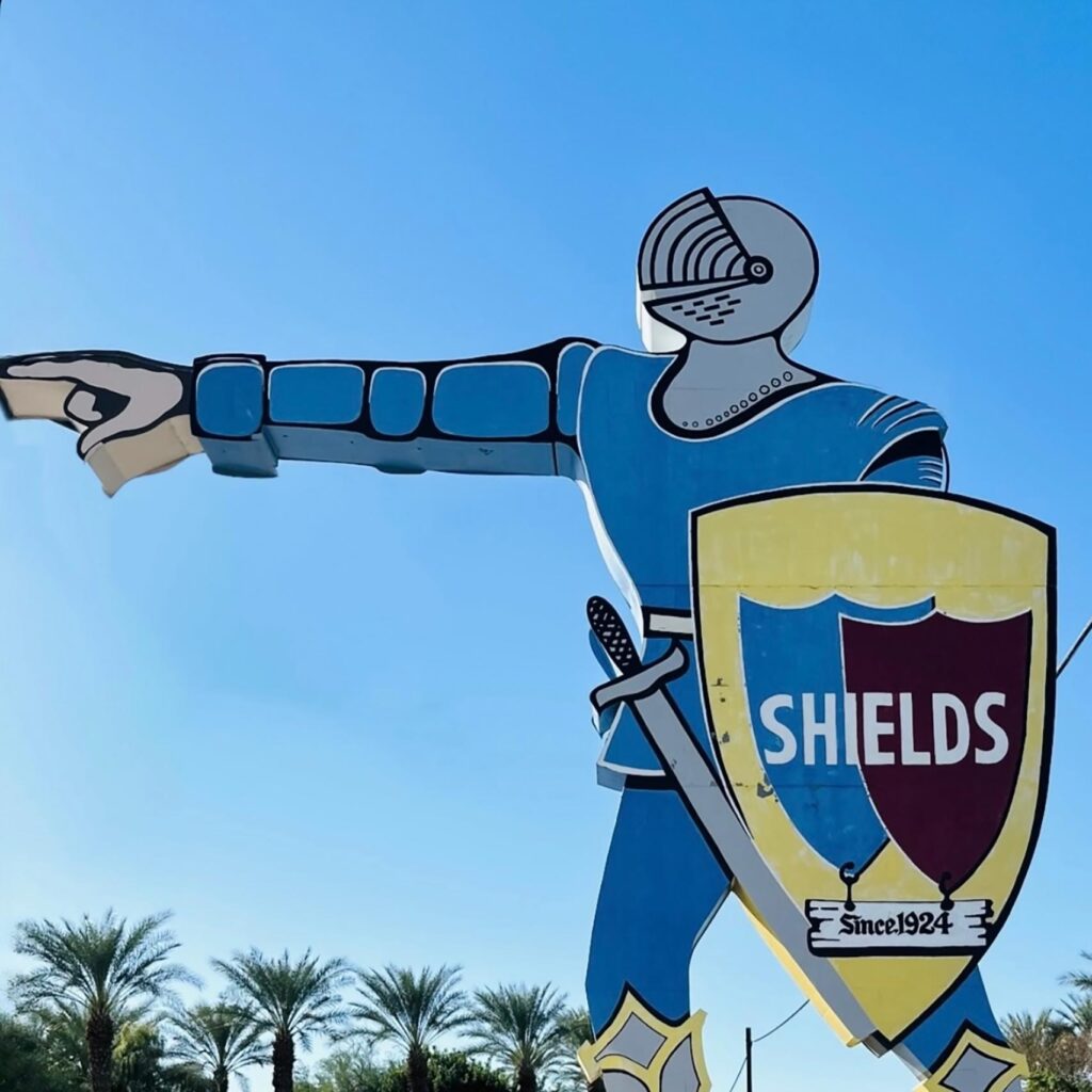A caricature sign of a knight in blue armour with a sword and shield. The shield is yellow and contains a smaller blue and black shield with the words SHIELDS in white. Beneath it hangs a sign that reads "Since 1924"