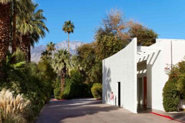 Entrance driveway to the Parker Palm Springs, white, mid-century facade, landscape palms and plans and the mountains and blue sky in the background. Outside the building, a man stands, wearing a pink jacket and white trousers