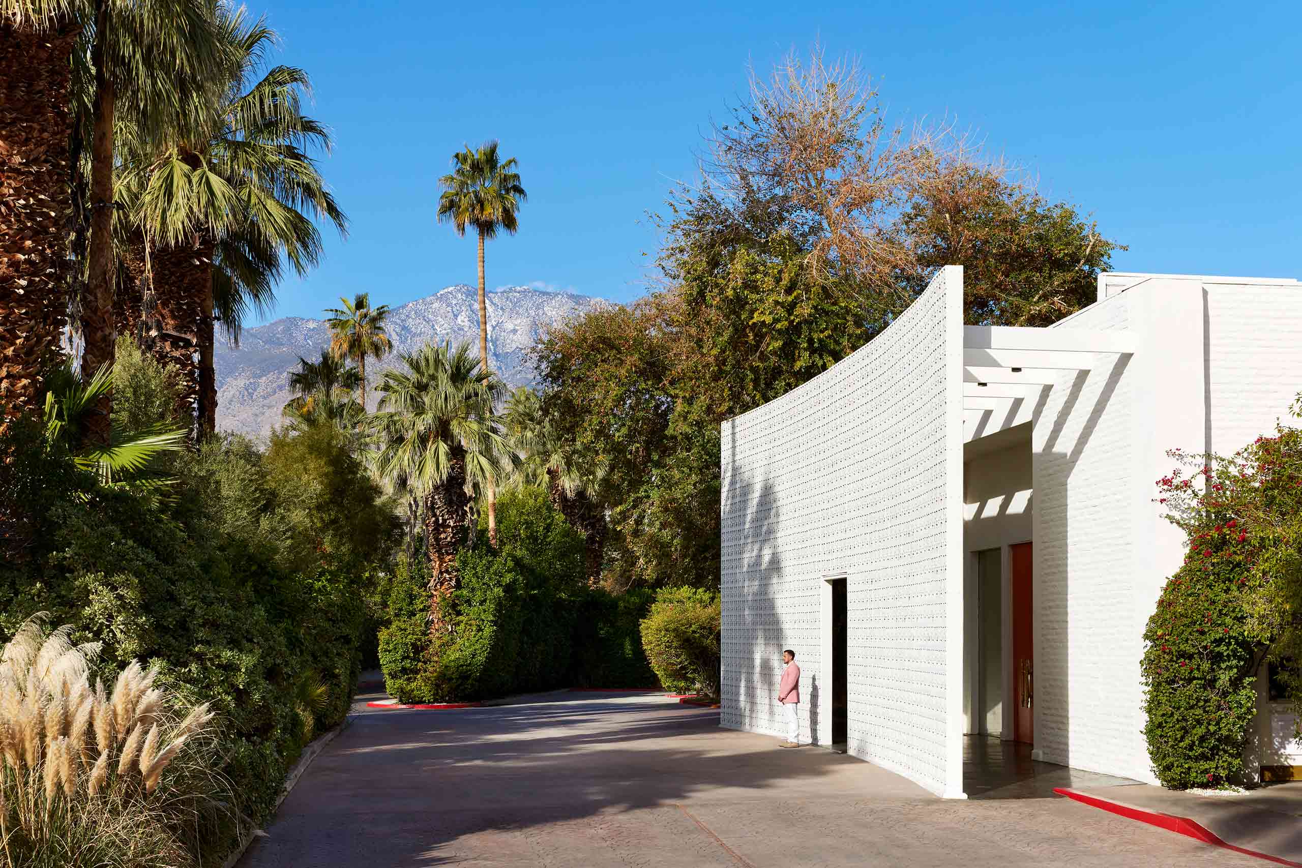 Entrance driveway to the Parker Palm Springs, white, mid-century facade, landscape palms and plans and the mountains and blue sky in the background. Outside the building, a man stands, wearing a pink jacket and white trousers