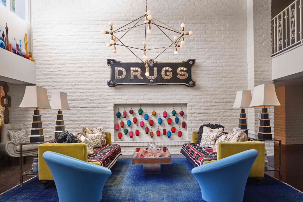 Lobby lounge of Parker Palm Springs with Jonathan Adler's eclectic furniture and esign pieces. At the centre of a white wall is a vintage drugstore sign that reads "DRUGS" – beneath it a collection of lanterns, sofas, armchairs, lamps and big blue rug