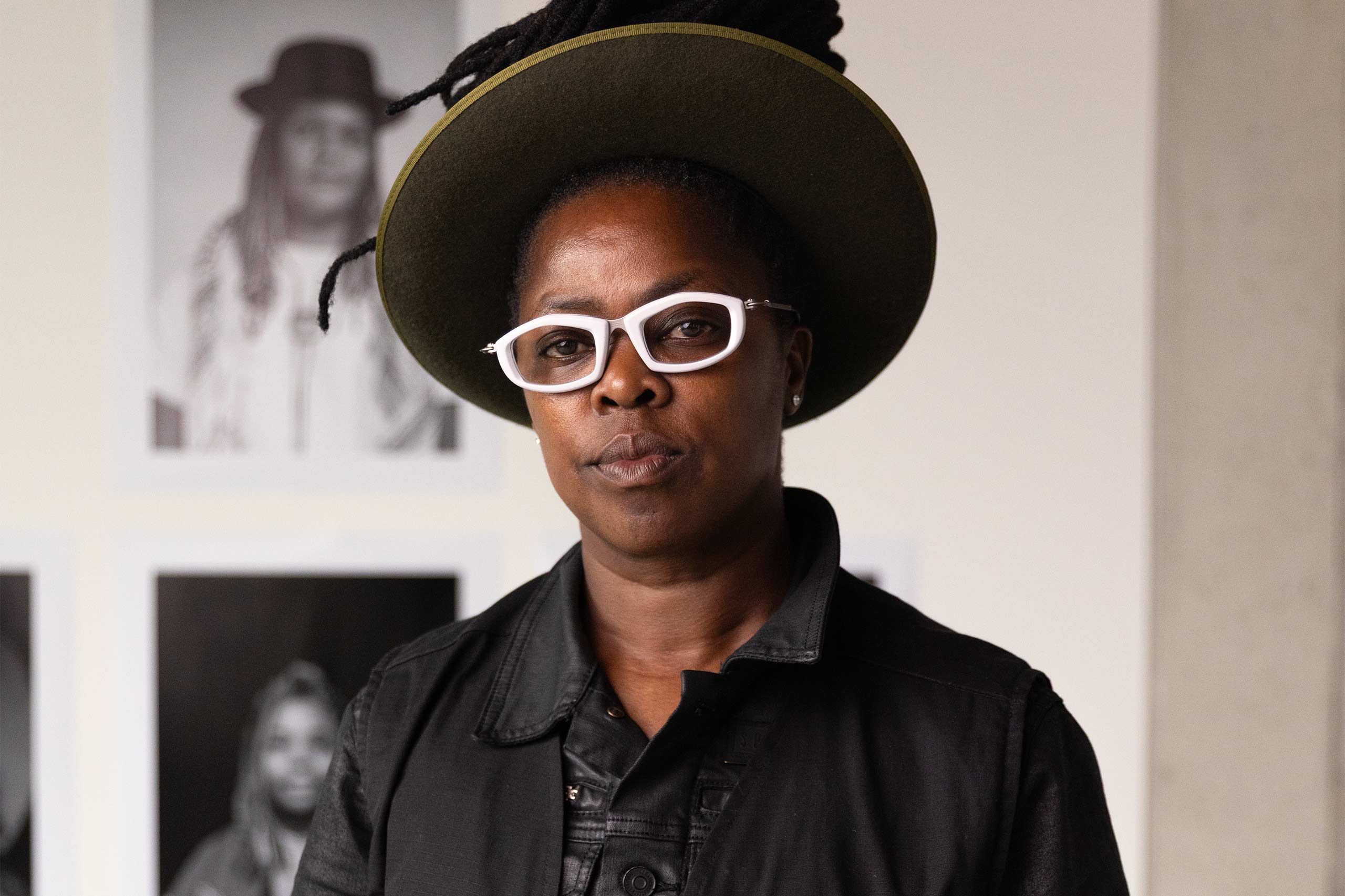 A portrait of Zanele Muholi at the QUEERCIRCLE in London. They wear a large green hat and chunky white glasses.