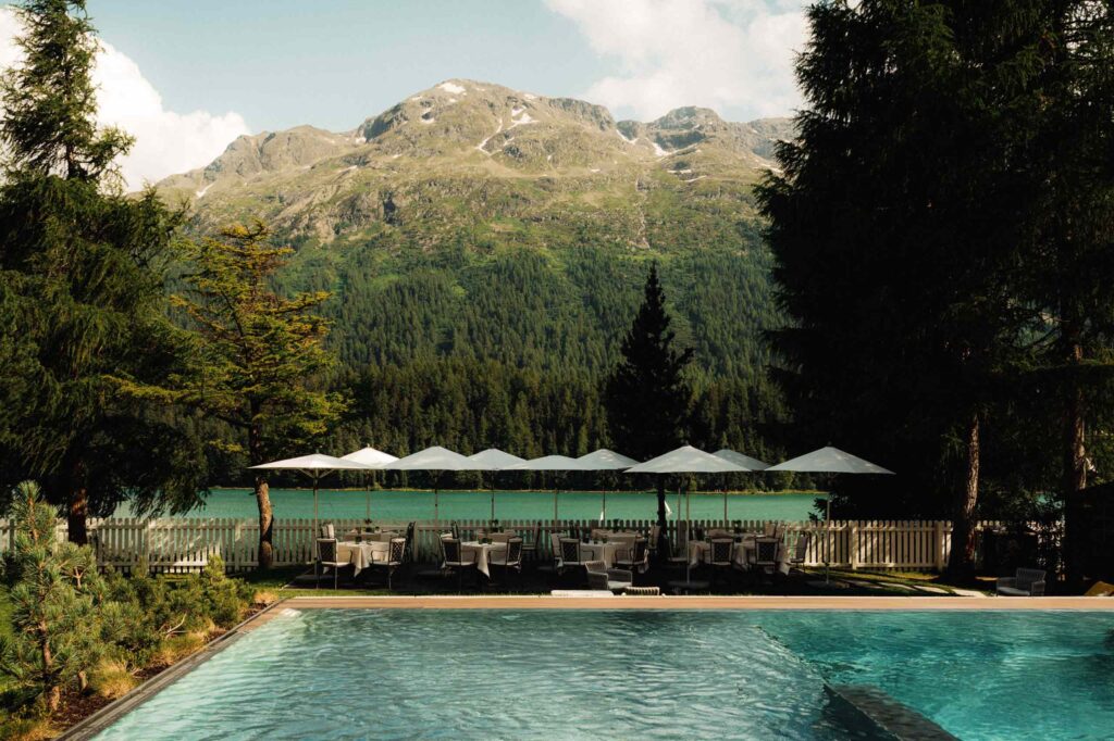 A pool with poolside loungers umbrellas in front of Lake St Moritz and the Alps