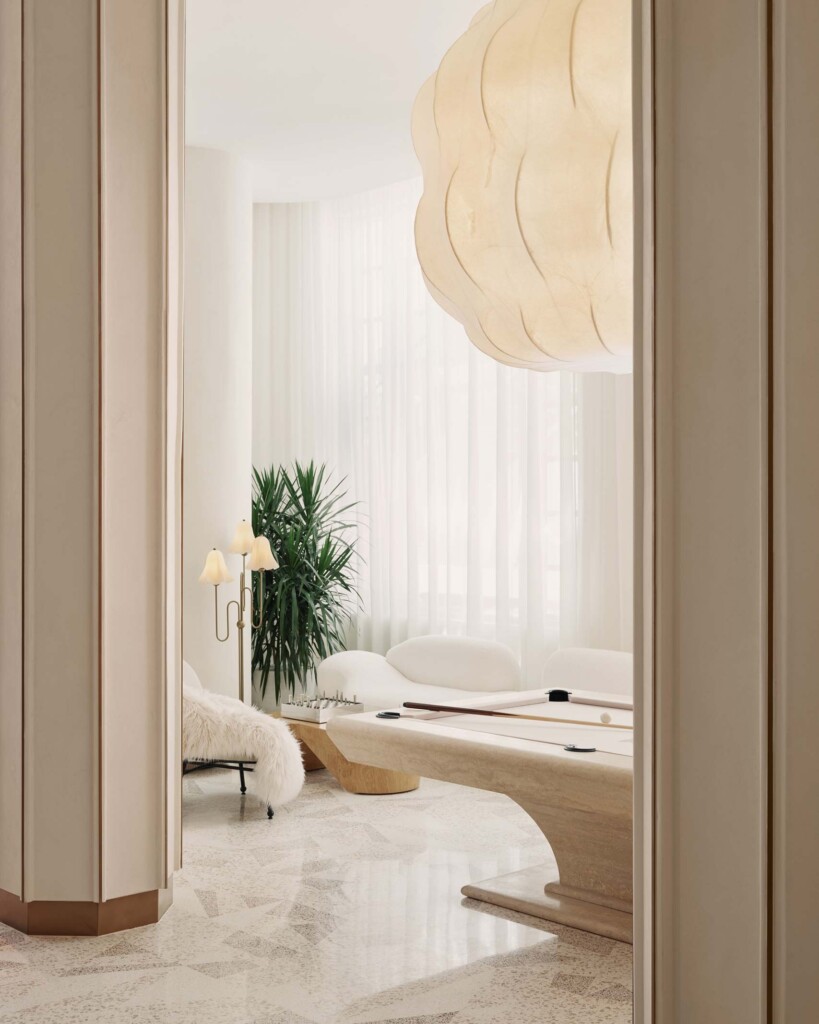 A white marble pool table sit below a large beige lantern and clean white interiors, flanked by 1947-revival hexagonal stone columns