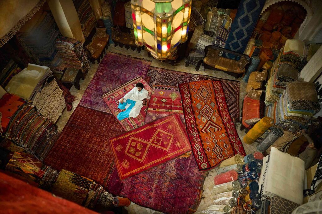 Amran Bennaji wearing a handwoven cotton tuxedo while reclining on red rugs beneath a Moroccan lamp in Marrakech, Morocco