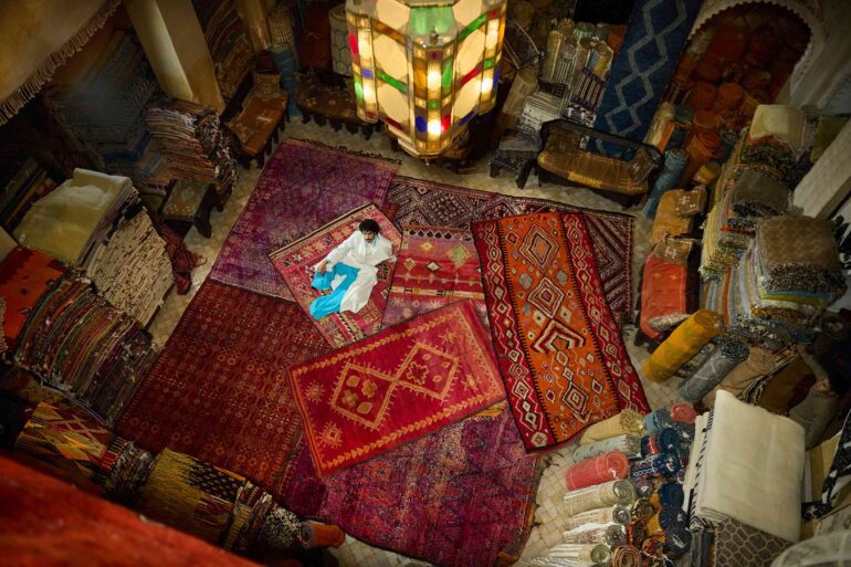 Amran Bennaji wearing a handwoven cotton tuxedo while reclining on red rugs beneath a Moroccan lamp in Marrakech, Morocco