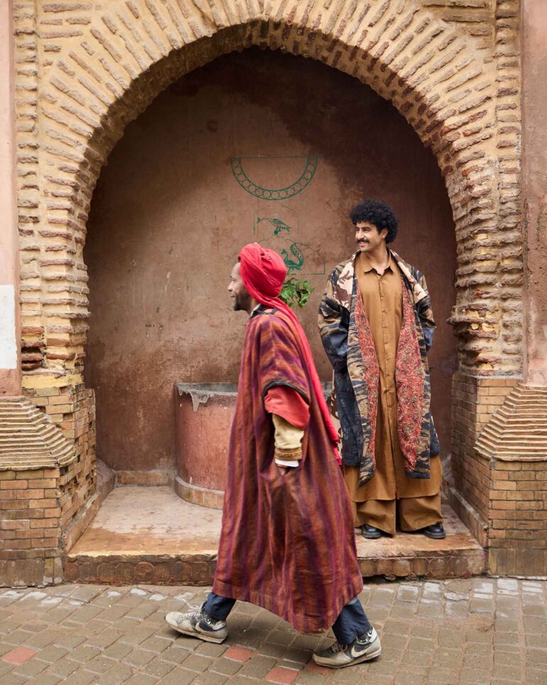 Amran Bennaji wearing a long, buttoned shirt while posing in front of a wooden doorway as a stranger walks past in the medina in Marrakech, Morocco